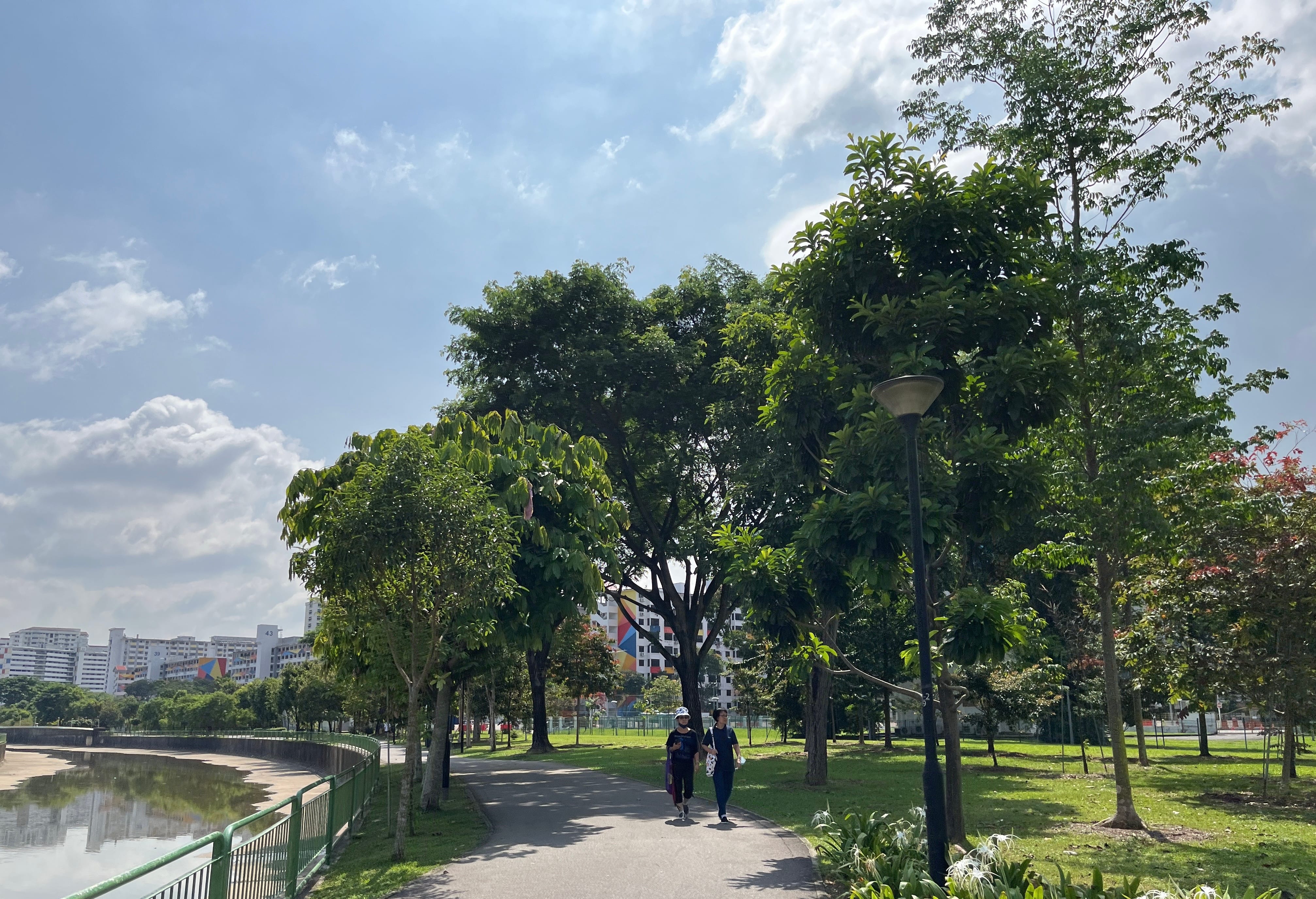 Two people are walking on a path in a park, surrounded by trees, with buildings visible in the background.